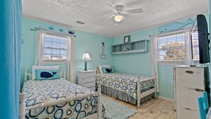 Une chambre avec deux lits, un ventilateur de plafond, une petite table avec une lampe et un réfrigérateur d’époque.