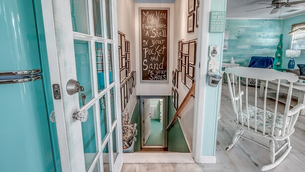 Un couloir étroit avec des portes de couleur turquoise, un escalier en bois et un petit fauteuil à bascule pour bébé. On peut voir un tableau noir sur lequel est écrit un message sur le thème de la famille.