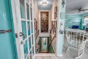 Un pasillo estrecho con puertas color turquesa, una escalera de madera y un balancín para bebé. Se ve una pizarra con un mensaje alusivo a la familia.