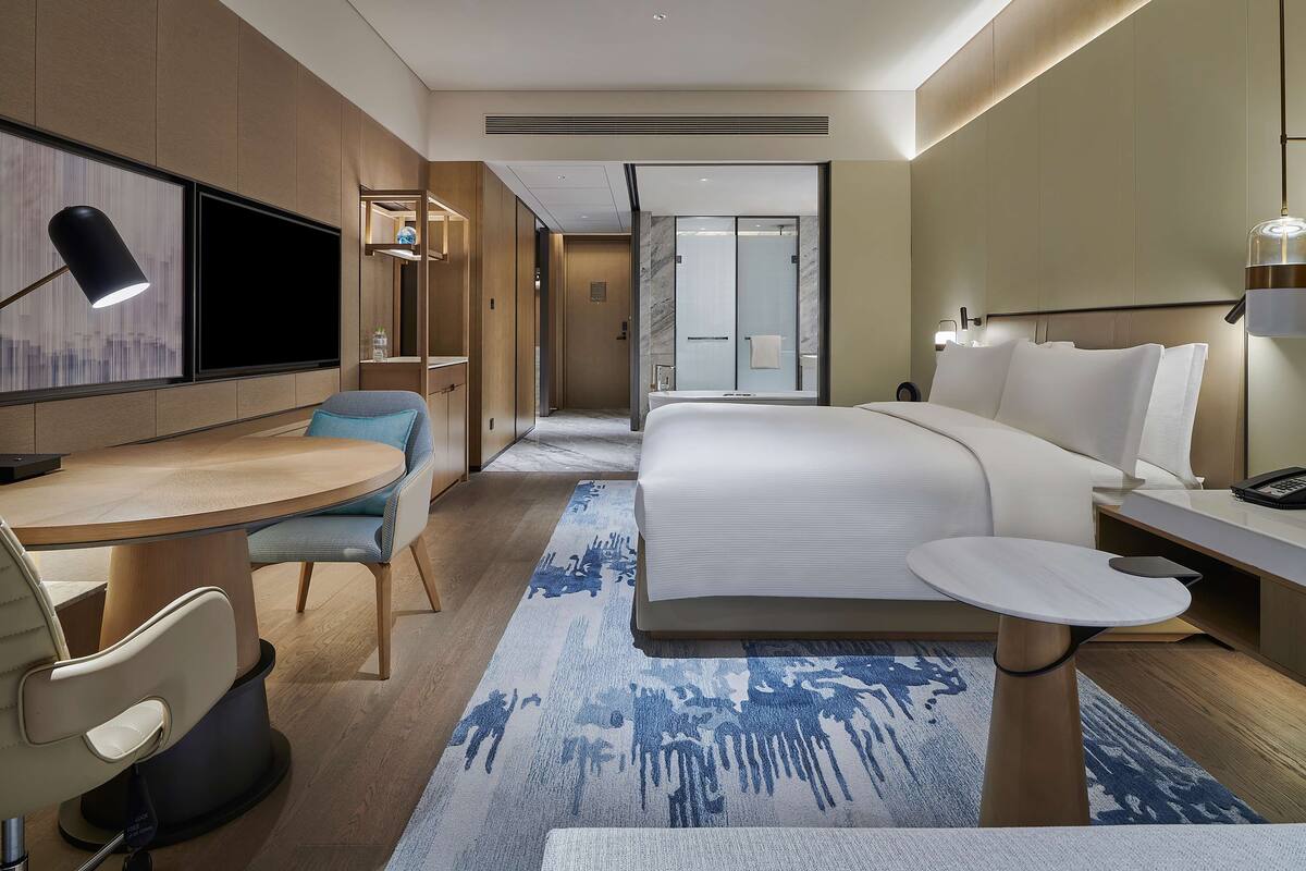 Une chambre d’hôtel moderne équipée d’un lit, d’une table de chevet, d’un bureau, d’une chaise et d’une grande fenêtre.