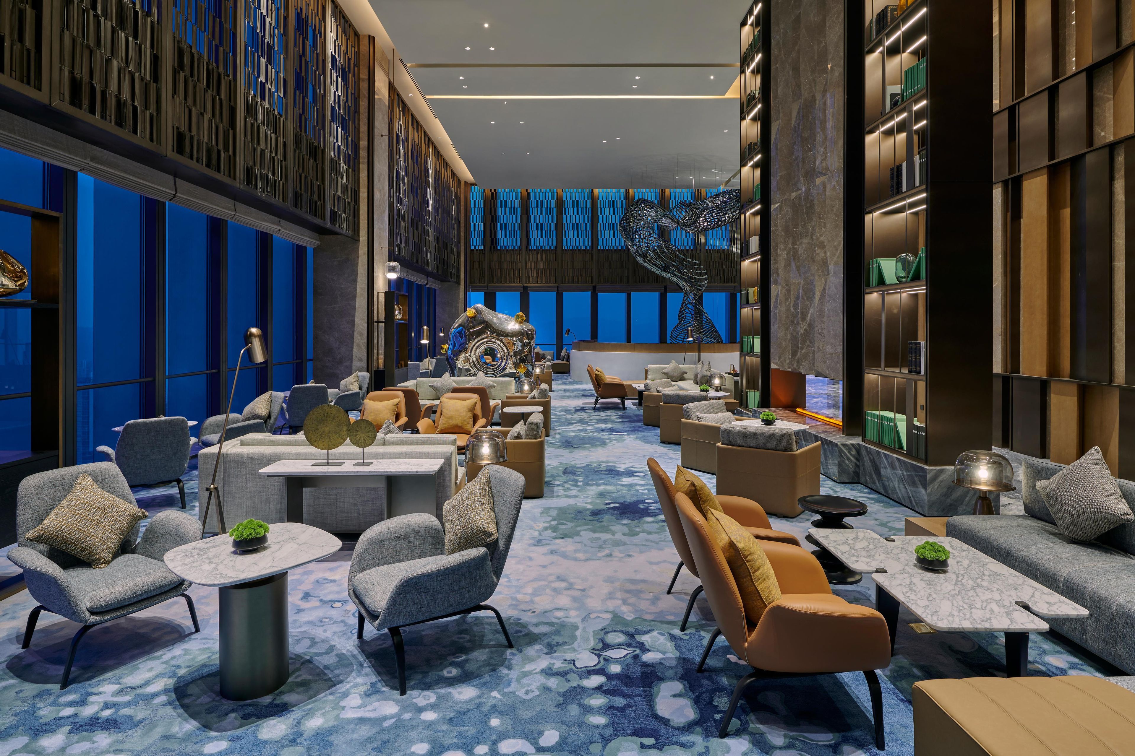 Un hall d’hôtel moderne, doté d’espaces aménagés avec des sièges, d’une sculpture centrale et d’une grande bibliothèque.