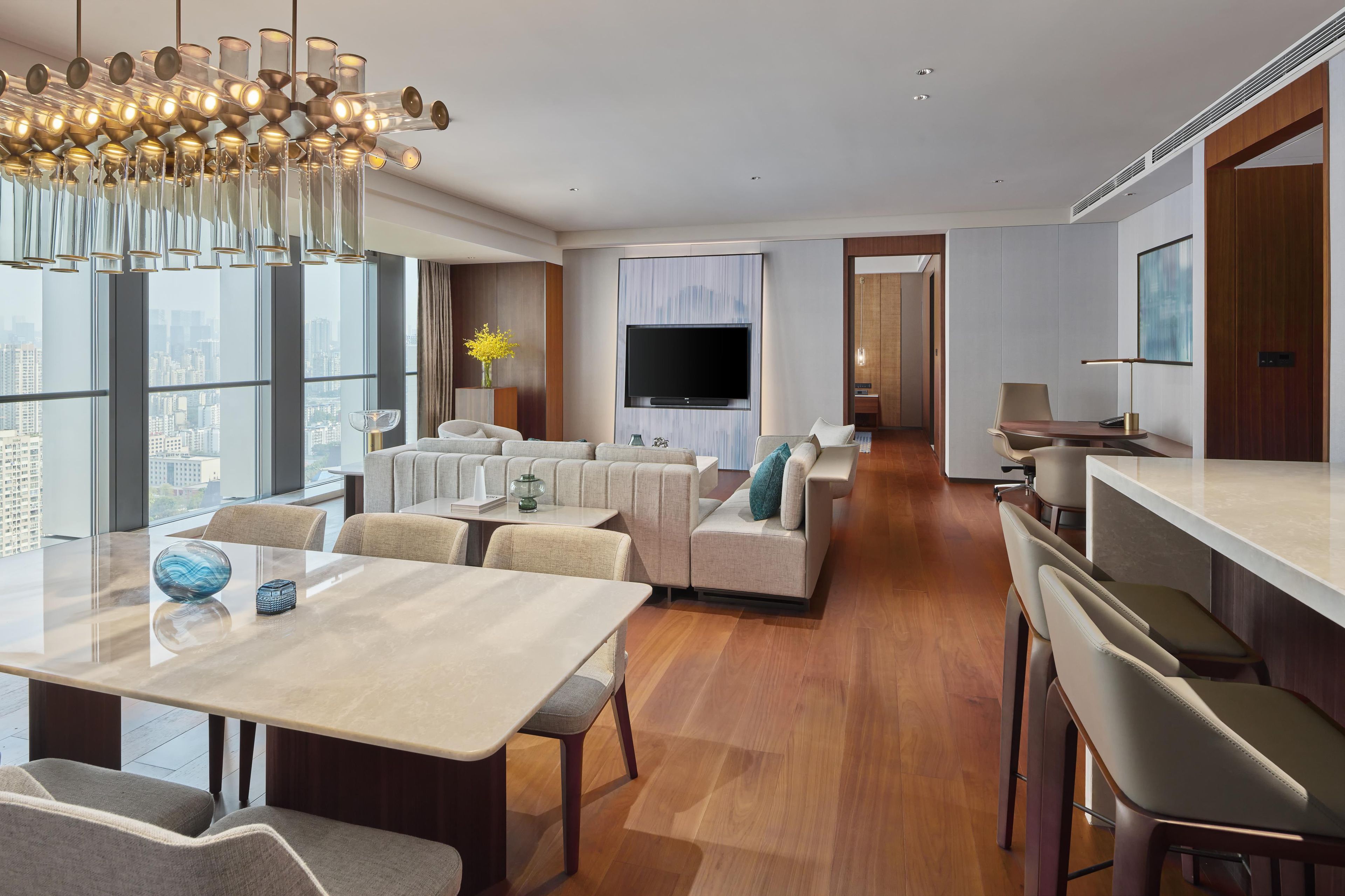 A modern hotel room with a dining area, a living room with sofas, a TV, and a city view.