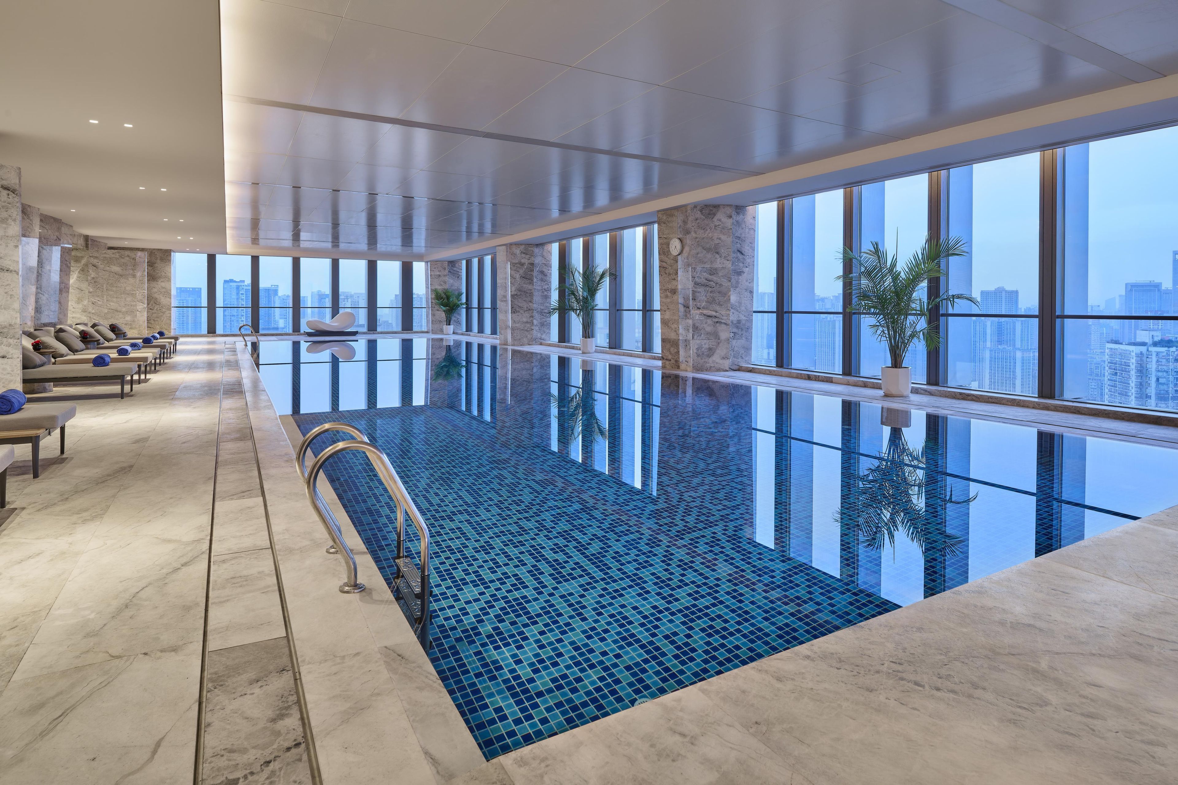 Una piscina interior moderna con vistas a la ciudad, suelo de mármol y zona de descanso.