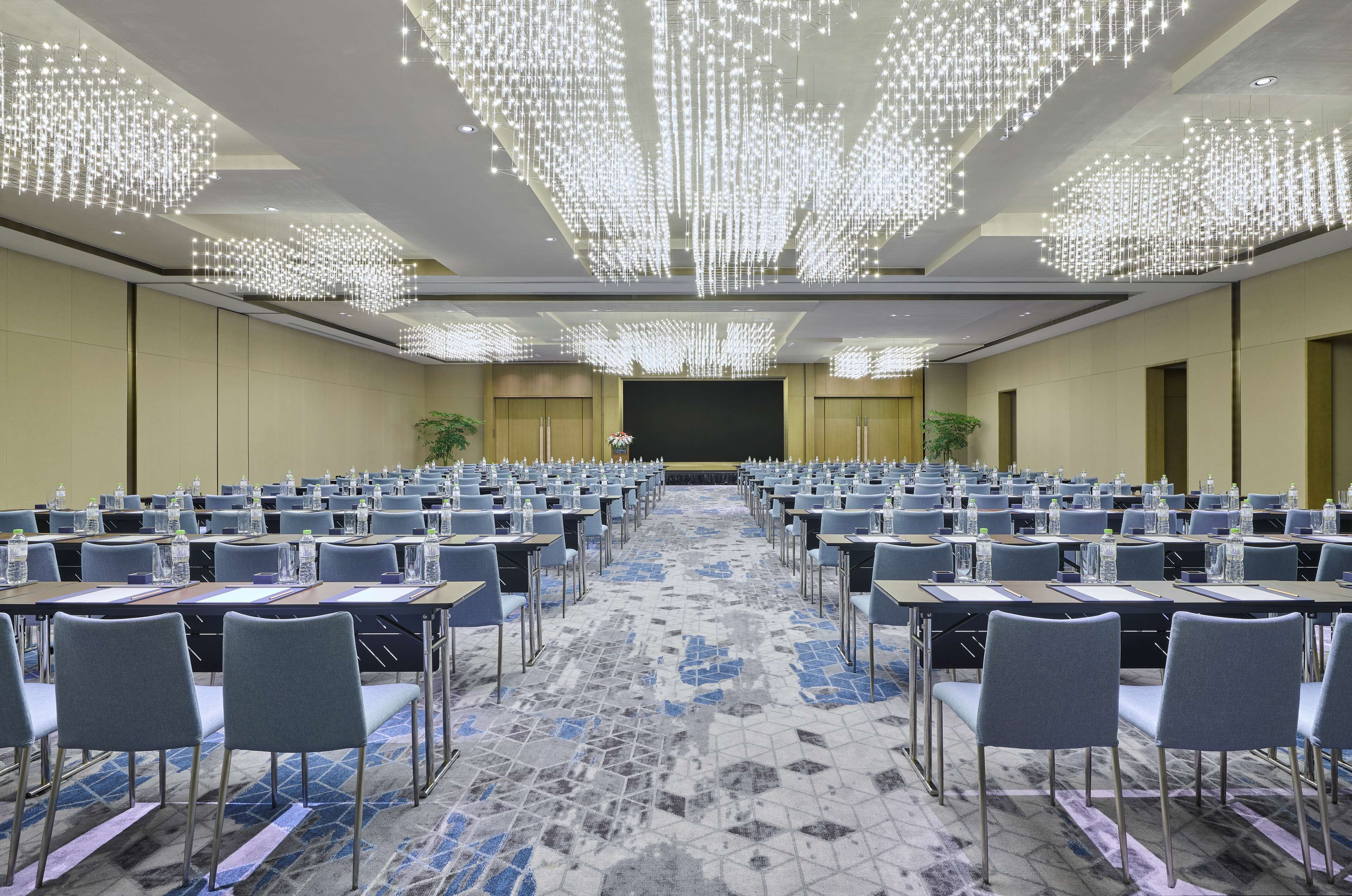 A conference room with rows of tables and chairs, a large screen at the front, and decorative ceiling lights.