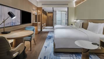 Une chambre d’hôtel moderne avec un grand lit, un bureau, une chaise et une petite table. Un téléviseur est fixé au mur, il y a un espace douche et la fenêtre offre une vue sur la ville.