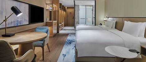 Ein modernes Hotelzimmer mit einem großen Bett, einem Schreibtisch, einem Stuhl und einem kleinen Tisch. An der Wand ist ein Fernseher befestigt, es gibt eine Duschkabine und einen Blick auf die Stadt durch das Fenster.