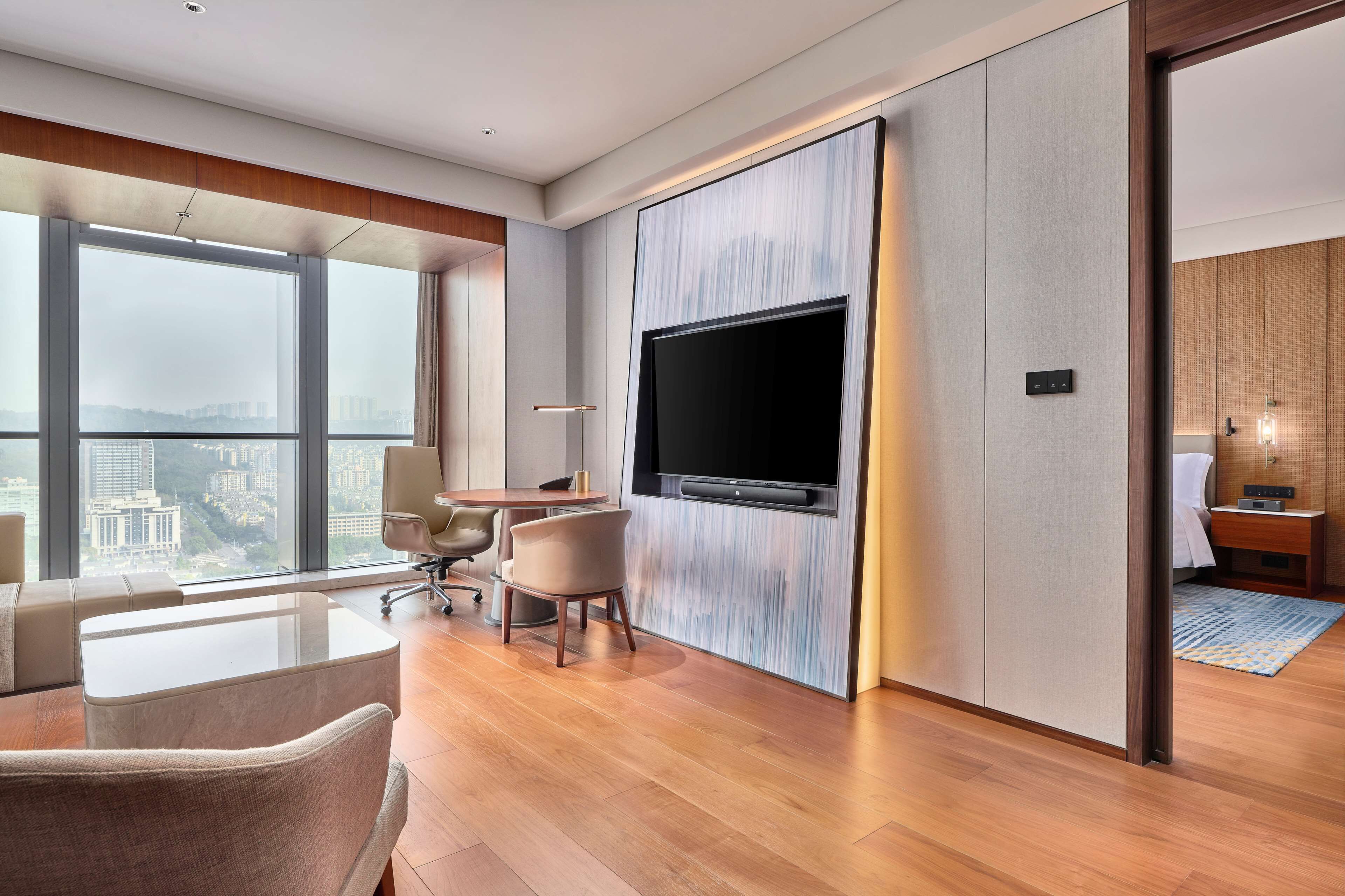Ein modernes Hotelzimmer mit großem Fenster, einem an der Wand befestigten Flachbildfernseher, Holzboden und einer Sitzecke mit Tisch und Stühlen.