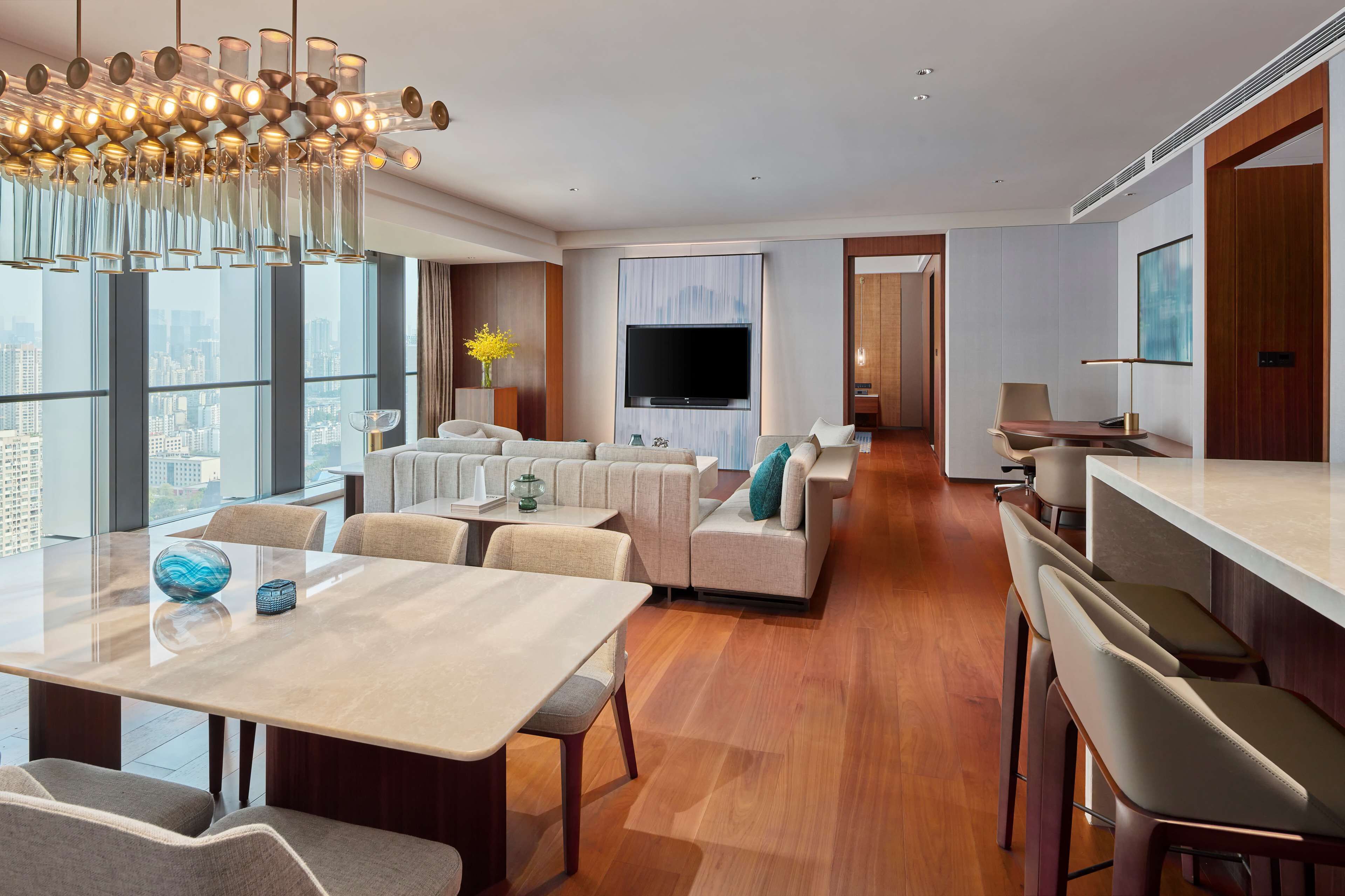 A modern hotel room with a large TV, comfortable seating, a dining area, and a view of the city.