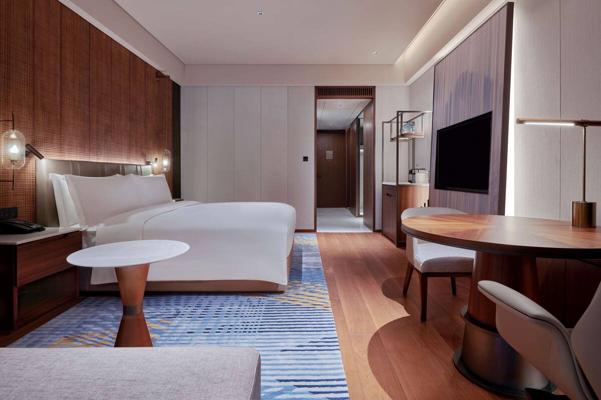 Une chambre d’hôtel moderne avec un grand lit, une petite table ronde, un bureau avec une chaise, une télévision et une salle de bain visible à travers une porte.