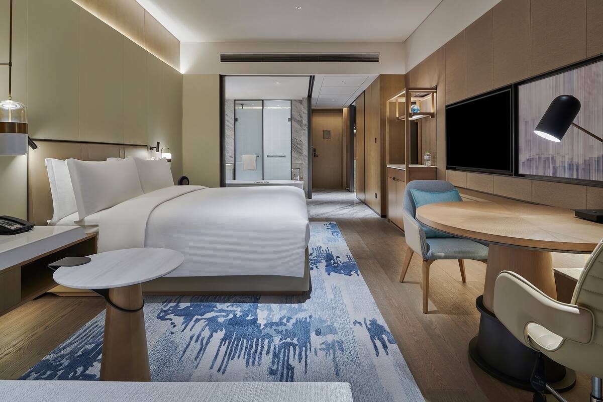 Une chambre d’hôtel moderne dotée d’un grand lit, d’un bureau avec une lampe, d’une télévision et d’une salle de bain visible à travers une porte en verre.