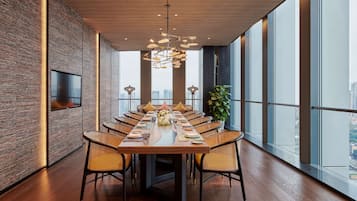 A modern dining area with a long table, wicker chairs, and a city view through large windows.