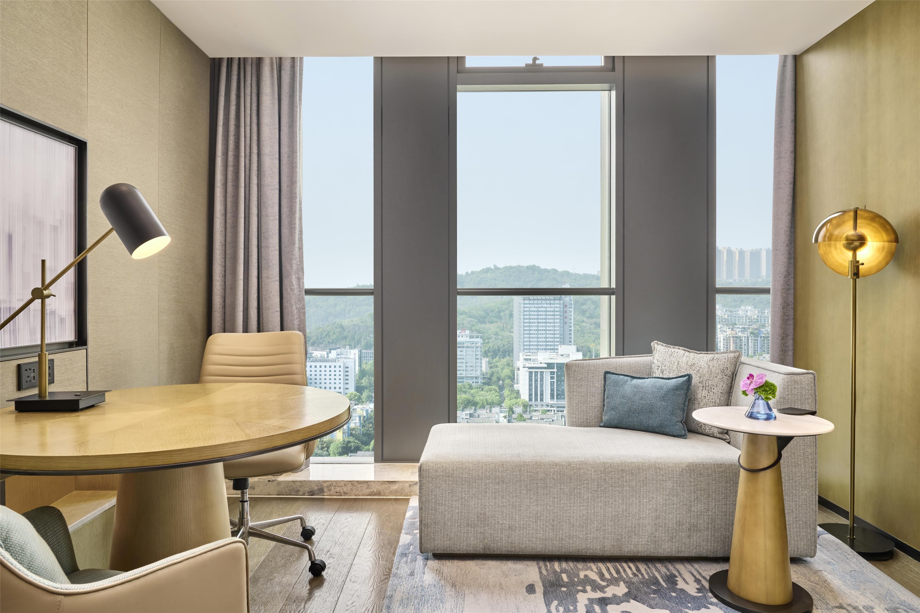 A modern hotel room with a large window offering a city view, a round dining table, a sofa, and a small side table with a lamp.