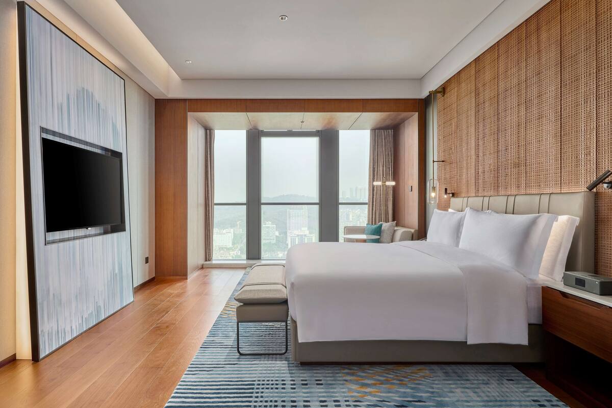 Une chambre d’hôtel moderne dotée d’un grand lit, d’un téléviseur à écran plat fixé au mur, d’une grande fenêtre offrant une vue sur la ville et d’une tête de lit en bois.