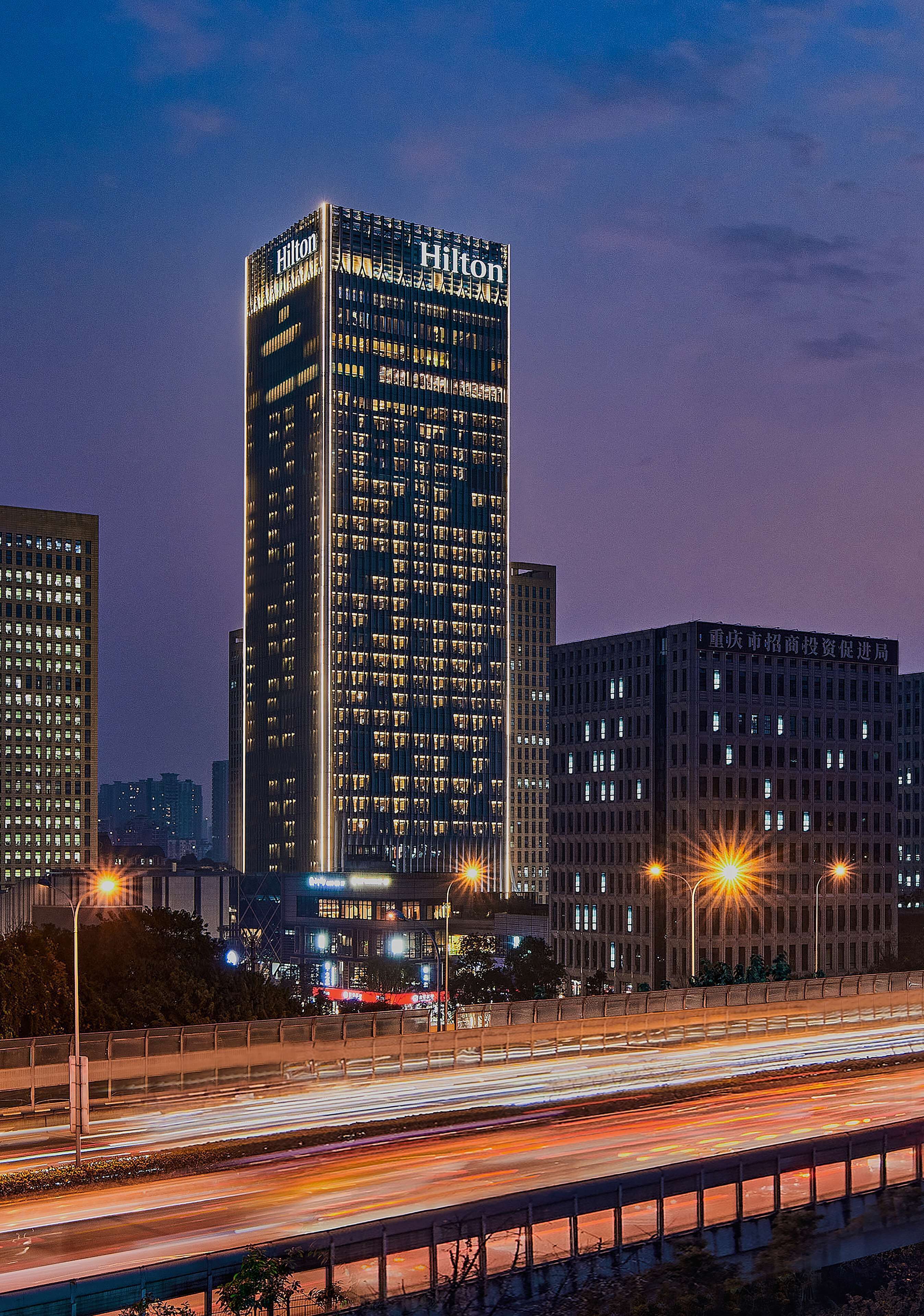 Un edificio alto e moderno con insegne illuminate, circondato da altri grattacieli e da un'affollata autostrada con scie luminose.