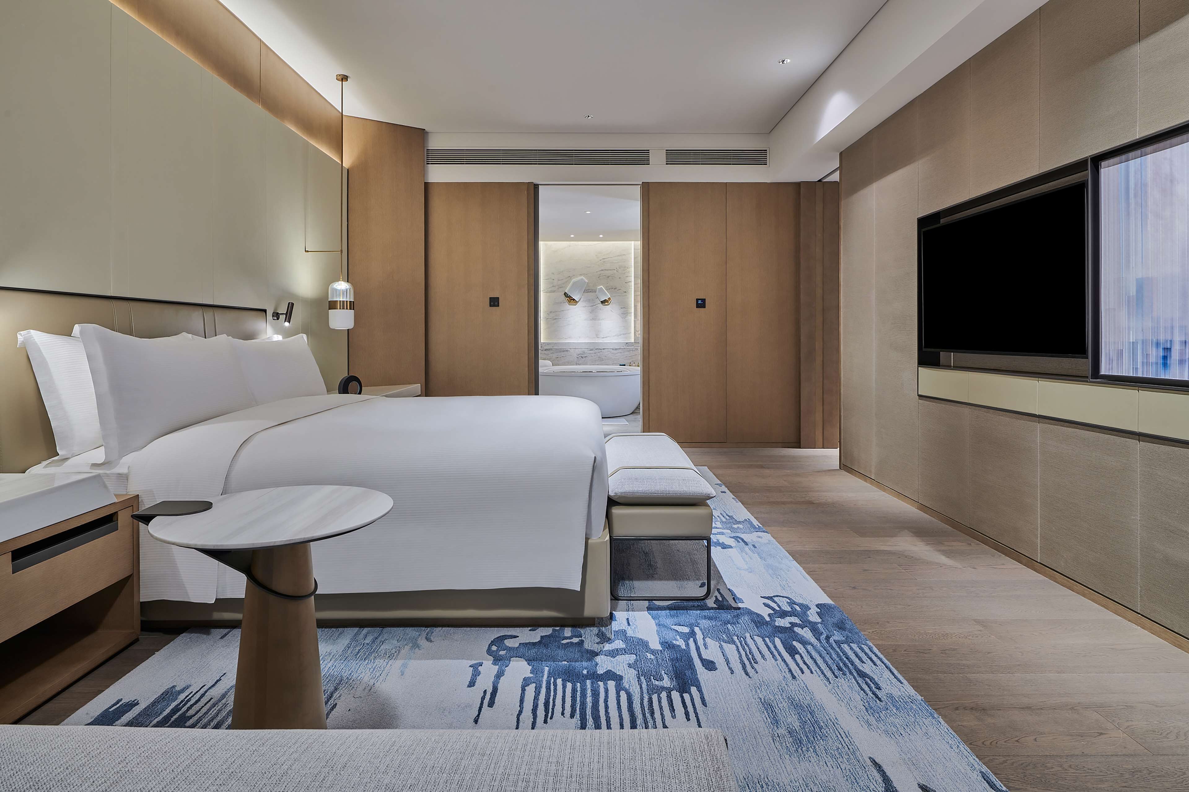 A modern hotel room with a large bed, bedside tables, a TV, and a view of a spa area.