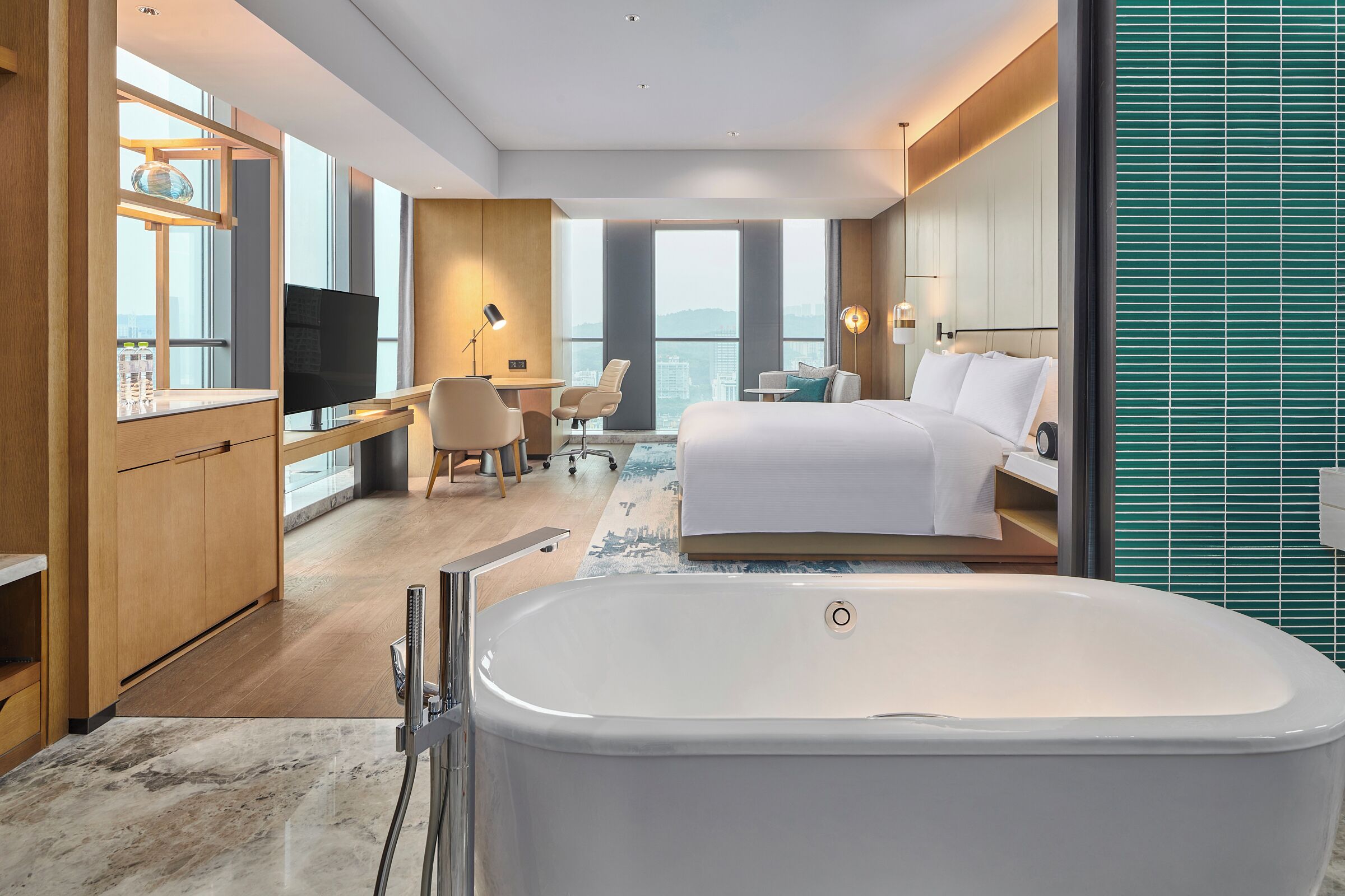 A modern hotel room with a large bed, a sitting area with a desk and chair, a bathtub, and a large window offering a city view.