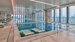 Une piscine intérieure moderne avec des parois en verre transparent, offrant une vue sur la ville, et un espace détente avec des chaises longues.