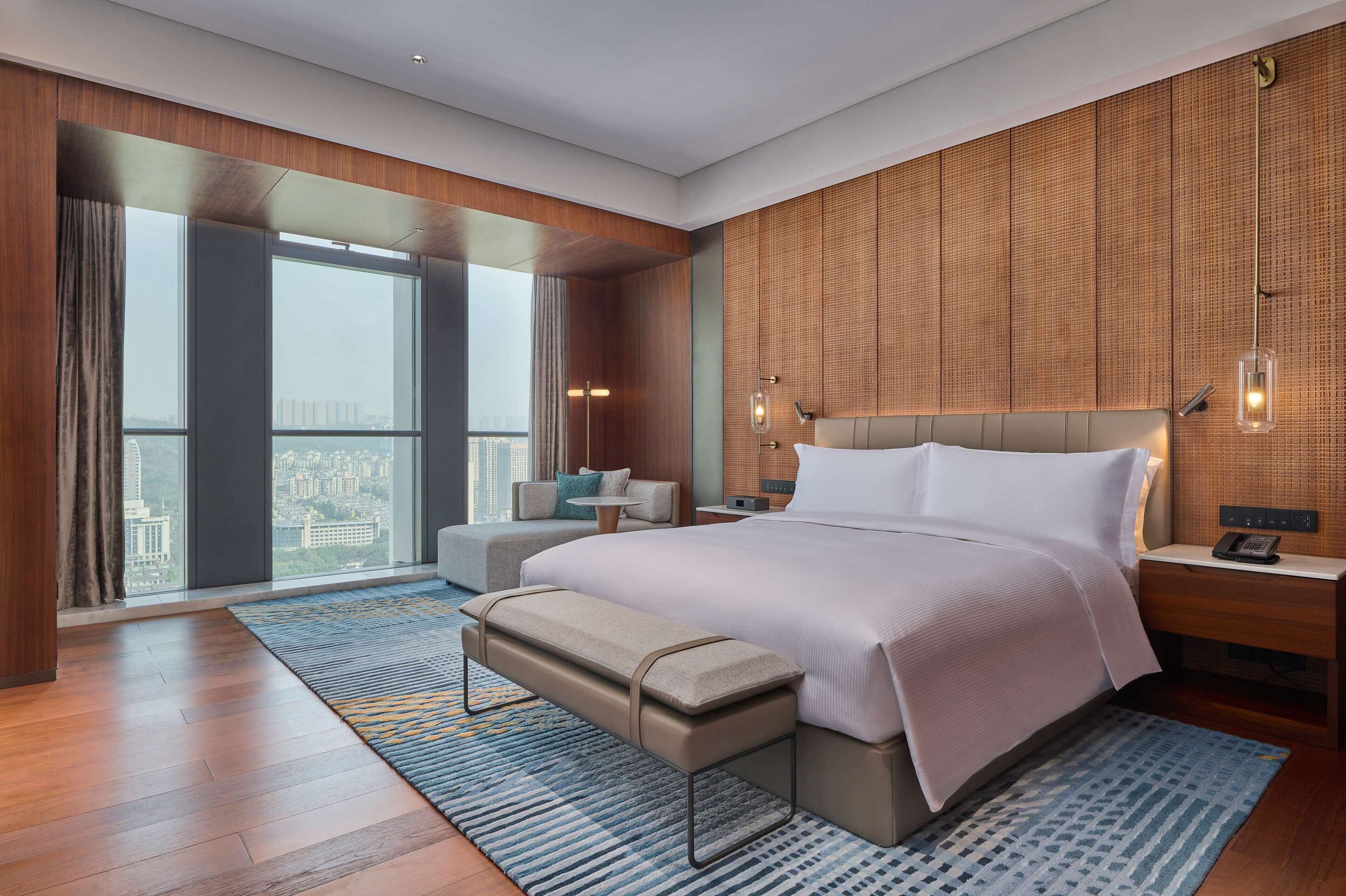 A modern hotel room with a large bed, a bench, a small table, and a view of the city through large windows.