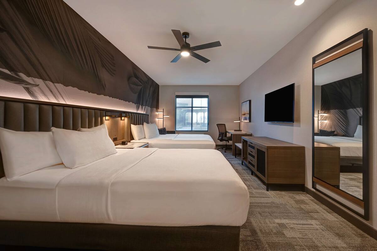A hotel room with two beds, a desk, a chair, a TV, a ceiling fan, and a large mirror.