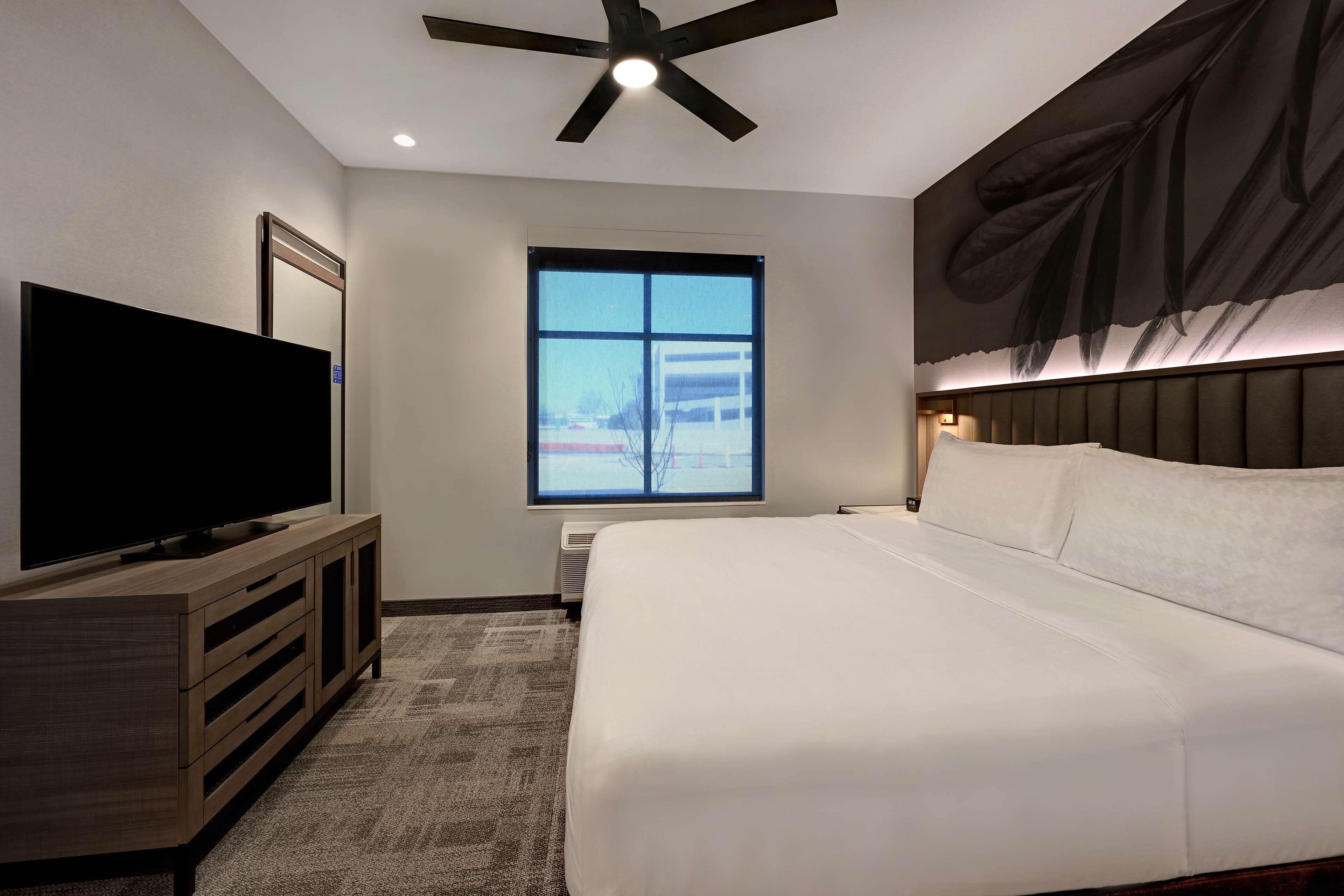 Una habitación de hotel moderna con una cama grande, un televisor de pantalla plana sobre un mueble de madera, un ventilador de techo, un ventanal con vistas a un edificio y una obra de arte colgada en la pared.
