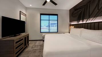 Una habitación de hotel moderna con una cama grande, un televisor de pantalla plana sobre un mueble de madera, un ventilador de techo, un ventanal con vistas a un edificio y una obra de arte colgada en la pared.