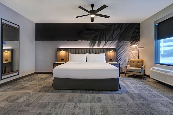 A hotel room with a large bed, a ceiling fan, a wall-mounted TV, a chair, and a window with a view.