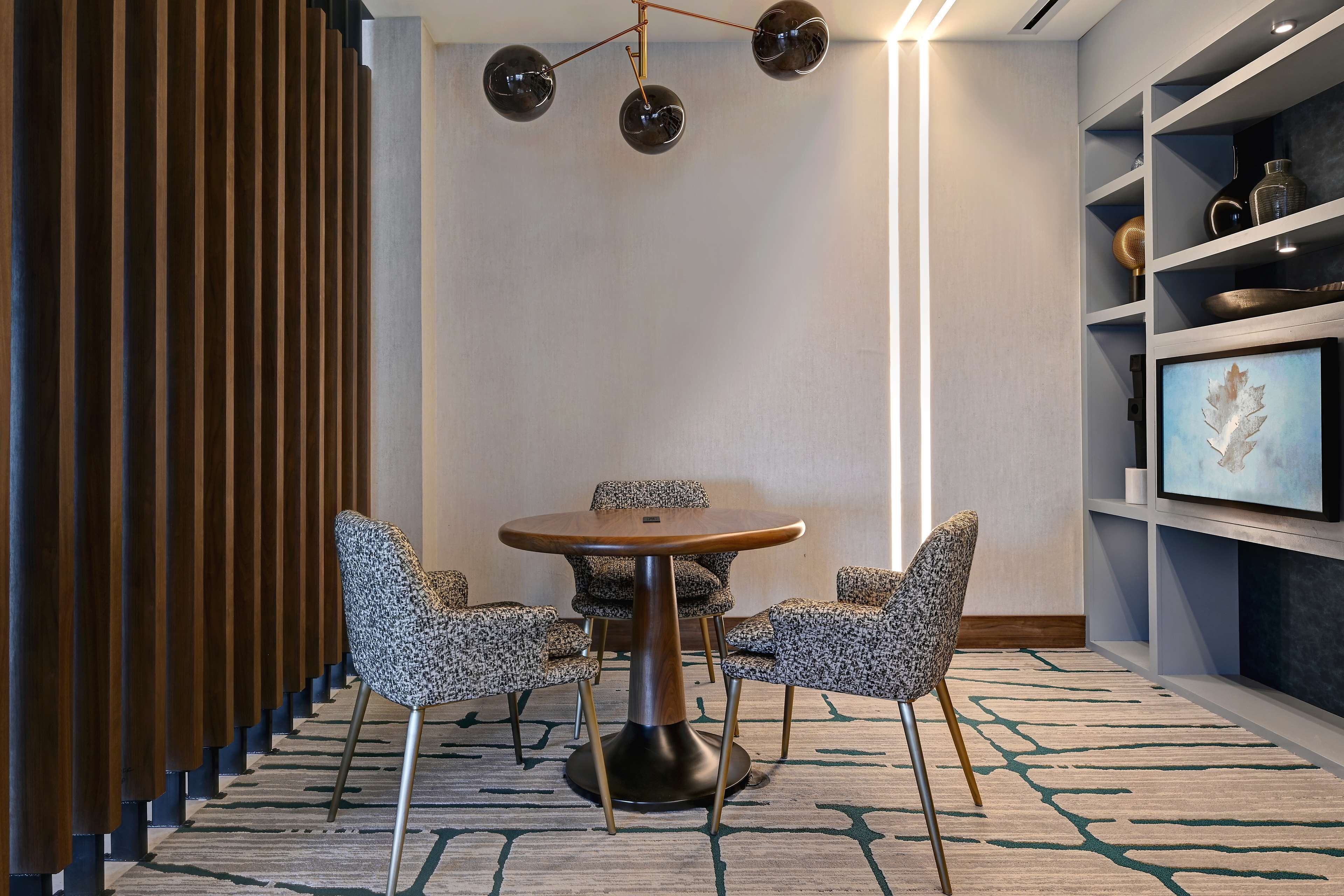 A conference room with a round table, four patterned chairs, a modern chandelier, and a bookshelf with decorative items.