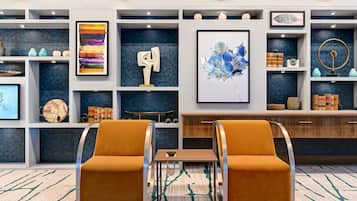 Un moderno vestíbulo de hotel con dos sillones naranjas, una mesa de centro de madera y una alfombra estampada. Estanterías exhiben diversos objetos decorativos y cuadros enmarcados.