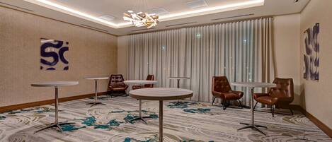 A hotel room with a patterned carpet, a chandelier, and a large window with curtains.