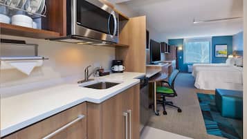 A modern hotel room with a kitchenette, a bed, a desk, and a large window.