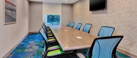 A conference room with a long table, blue chairs, a mounted TV, and a colorful carpet.