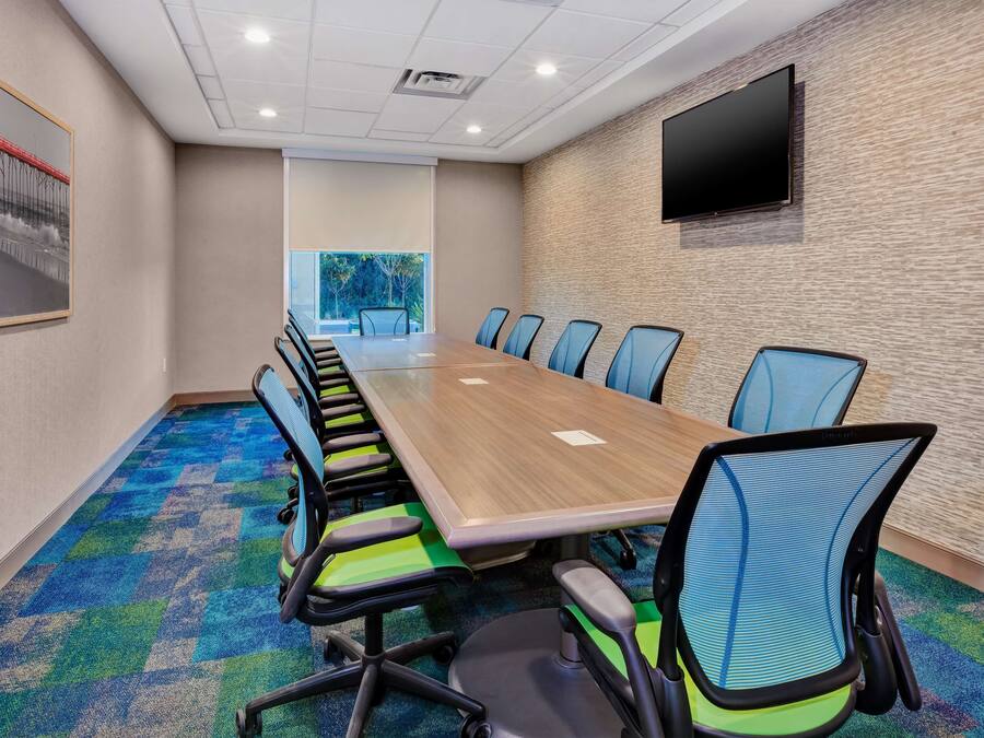 Een vergaderzaal met een lange tafel, blauwe stoelen, een wandgemonteerde televisie en een kleurrijk tapijt.