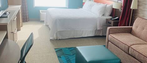 Una camera d'albergo con un letto, una scrivania con una sedia, un divano, un poggiapiedi e una finestra con tende.