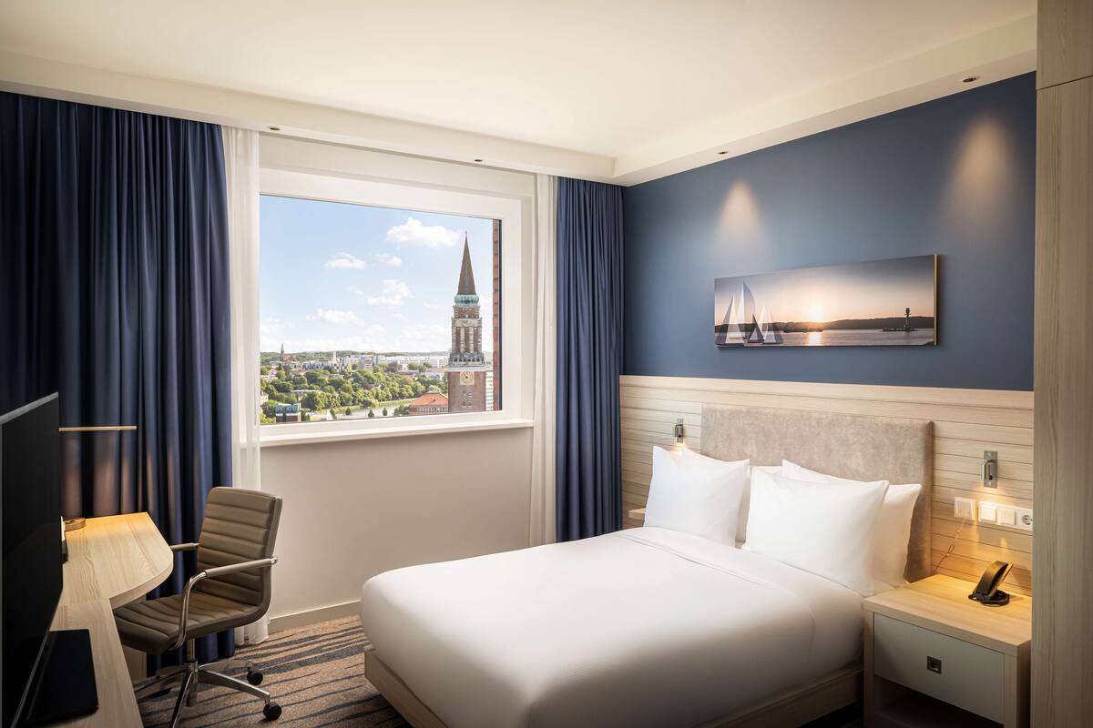 A hotel room with a bed, a desk with a lamp, a chair, a nightstand with a phone, and a window with a cityscape view.