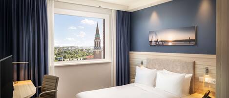 Habitación de hotel con cama, escritorio con lámpara, silla, mesita de noche con teléfono y ventana con vista panorámica de la ciudad.