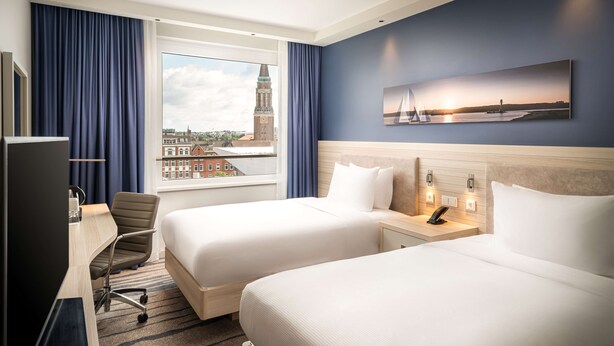 Quarto de hotel com duas camas, uma escrivaninha e uma cadeira. Há uma janela grande com vista para um horizonte urbano e um campanário.