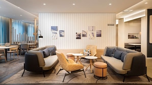 Um hall de hotel moderno com área de estar, incluindo sofás e cadeiras, uma mesa de centro e uma pequena mesa redonda. Há obras de arte penduradas na parede e uma placa indicando a área de café da manhã.