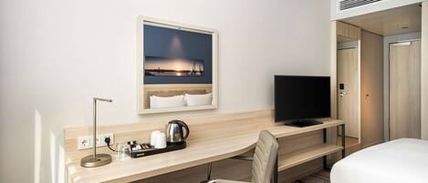 Una habitación de hotel con cama, escritorio con silla, televisor, cafetera y un cuadro de una puesta de sol sobre un puente.