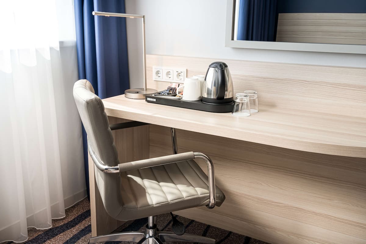 A hotel room with a wooden desk, a modern chair, a kettle, glasses, and a power outlet.