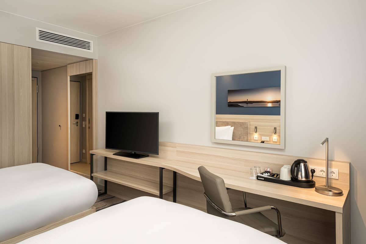 A hotel room with a bed, a desk with a TV, a chair, a bedside table with a kettle, and a framed picture on the wall.
