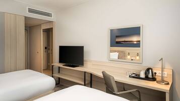 Ein Hotelzimmer mit Bett, Schreibtisch mit Fernseher, Stuhl, Nachttisch mit Wasserkocher und einem gerahmten Bild an der Wand.