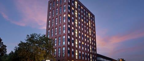 A tall, modern hotel with a prominent "Hampton" sign at the top, featuring a grid of windows and a flat roof.