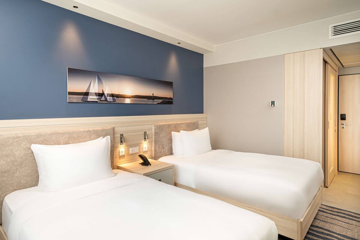 A hotel room with two beds, a wall-mounted painting of a sunset, and a wooden wardrobe.