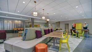 A spacious indoor area with a long table, multiple chairs, a sofa with cushions, and a seating area with a counter in the background. - Home2 Suites by Hilton Raleigh North I-540 (Raleigh)