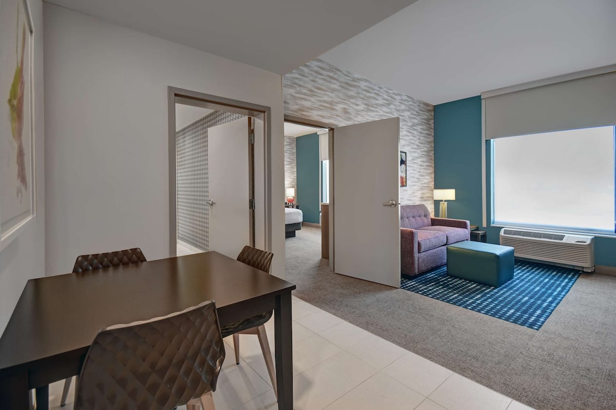 A modern hotel room with a dining area, a living space with a sofa and ottoman, and a large window providing natural light.