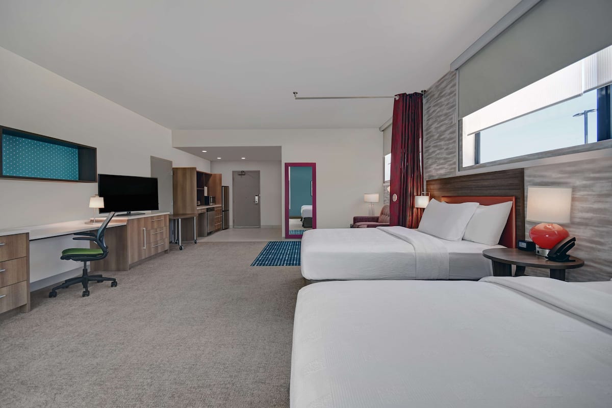 A hotel room with two beds, a desk with a chair, a TV, and a bathroom visible through an open door.