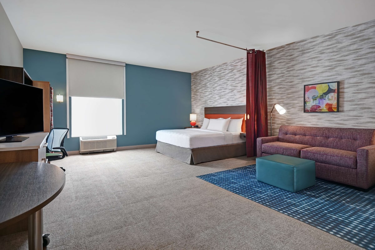 A hotel room with a bed, a sofa, a desk with a TV, and a large window with blinds.