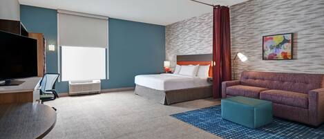 A hotel room with a bed, a sofa, a desk with a TV, and a large window with blinds.