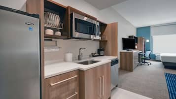 Una cocina moderna con refrigerador, microondas y lavamanos. Se observa un armario de madera con vajilla y un pequeño espacio de trabajo con escritorio y silla.