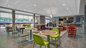 A modern indoor space with multiple seating areas, including round tables and chairs, and a counter with a menu board. - Home2 Suites by Hilton Raleigh North I-540 (Raleigh)